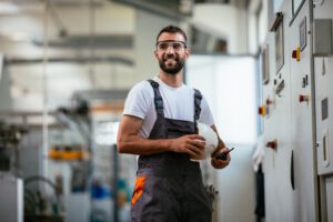 Industriemechaniker in Arbeitskleidung und mit Schutzbrille macht eine kleine Pause. Er hält in seinen Händen ein Becher mit Kaffee, ein kleines Funkgerät und Schutzhelm