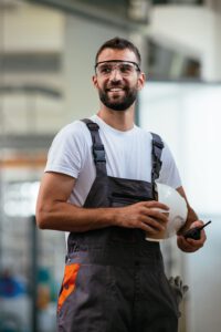 Industriemechaniker in Arbeitskleidung und mit Schutzbrille macht kurze Pause. Er hält in seinen Händen ein Becher mit Kaffee, ein kleines Funkgerät und Schutzhelm